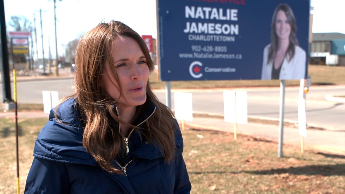 Woman wears blue coat and stands in front of election sign with her picture on it. 
