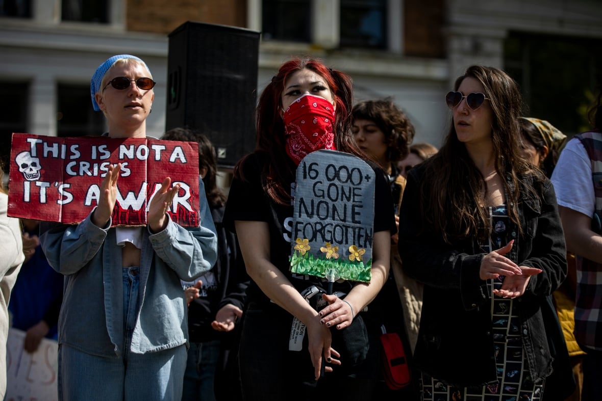 Three people, including a woman wearing a red mask, stand side-by-side, holding signs.