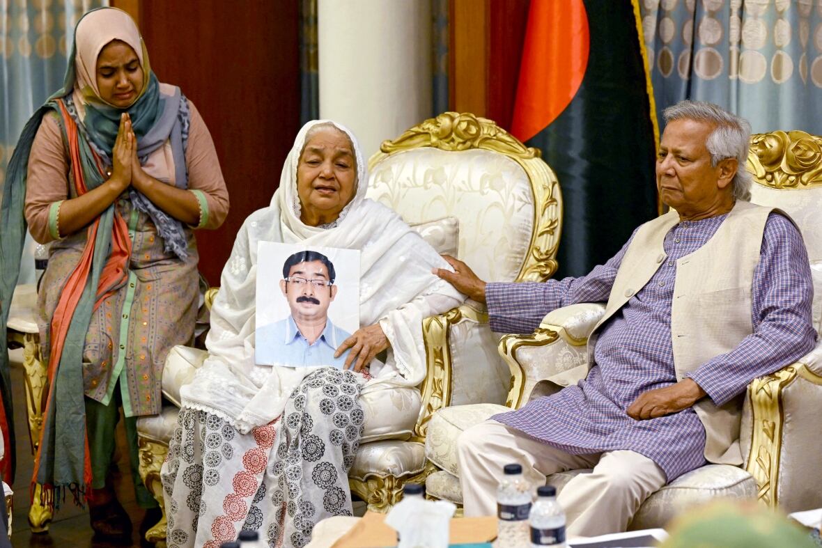 A woman in white, holding a photograph of a man with a moustache, sits next to a man with grey hair who is touching her arm while another woman looks on and holds her hands in prayer. 