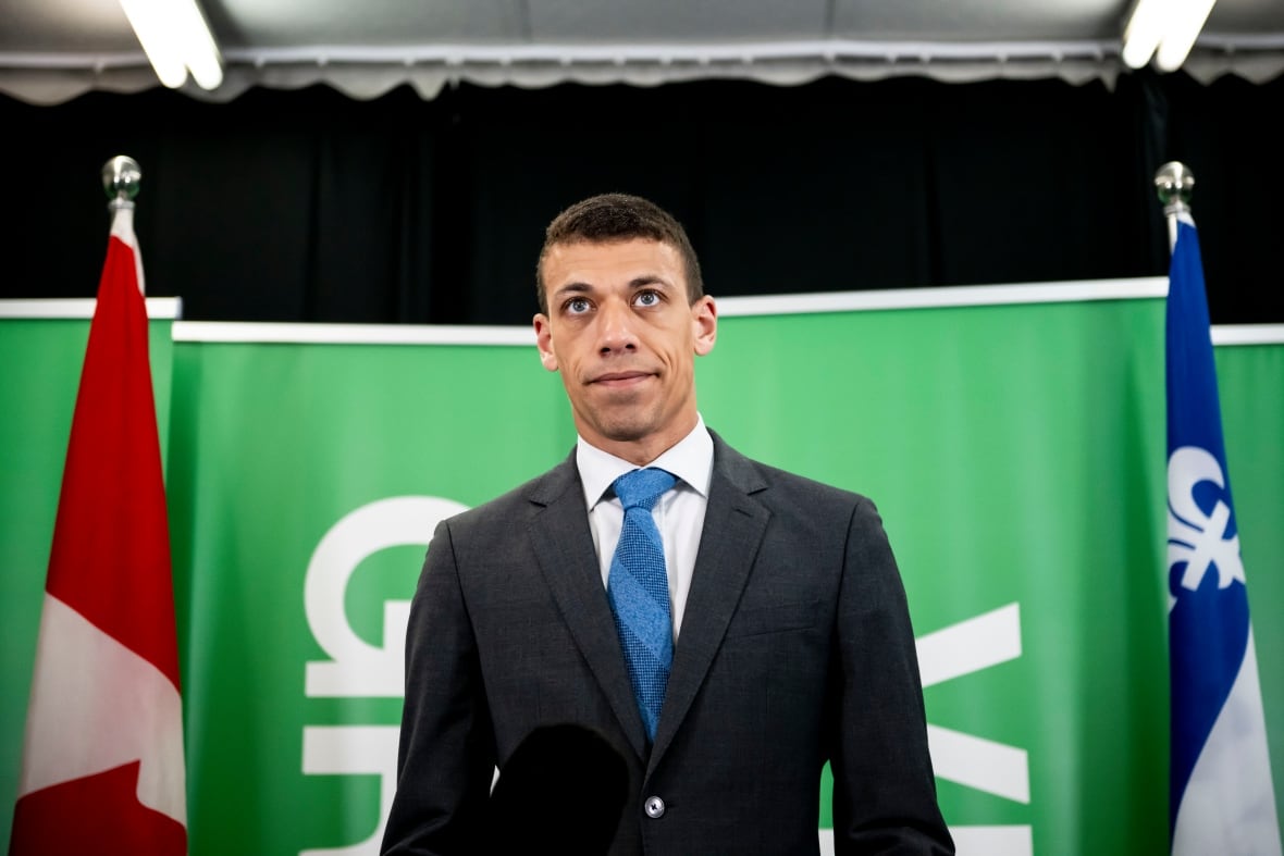 Um homem de terno fica em frente ao fundo verde.