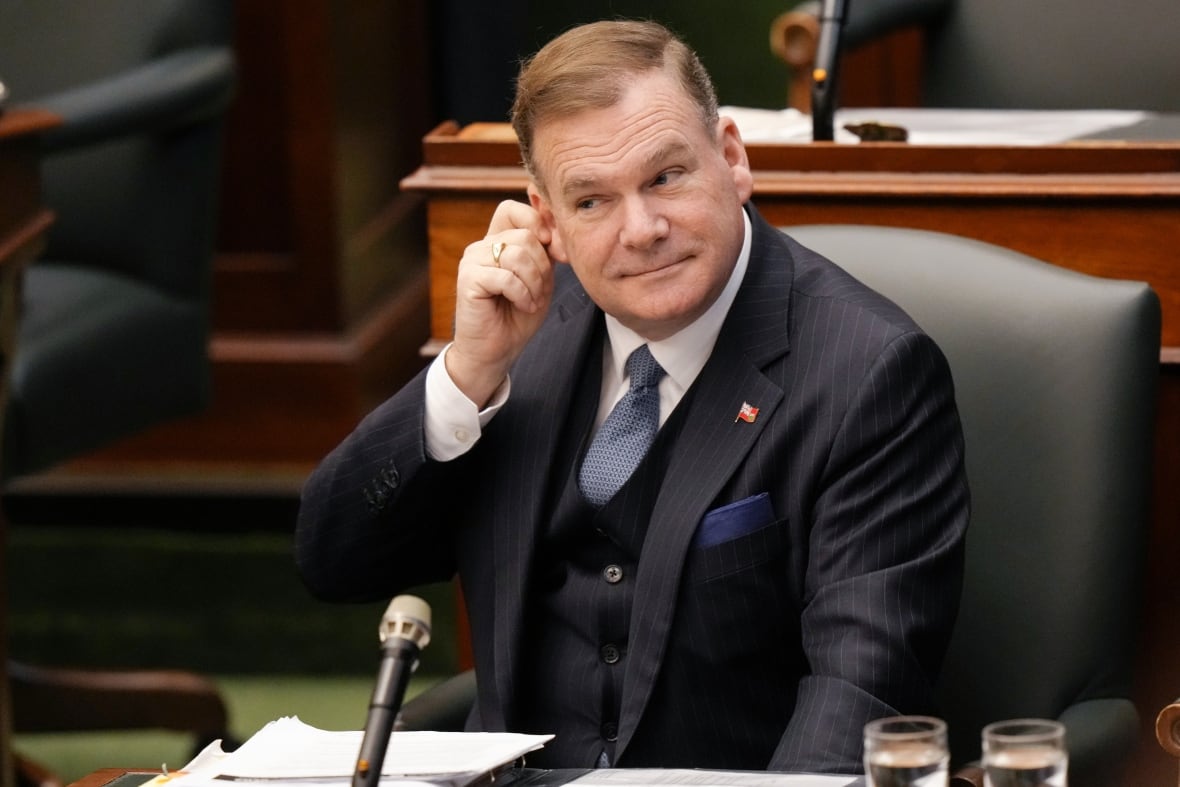 Man in a suit sits at Ontario legislature, smiling and touching ear