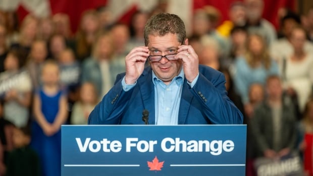 A man in a blue suit adjusts his glasses while standing onstage behind a podium that reads 'Vote for Change' in both English and French.