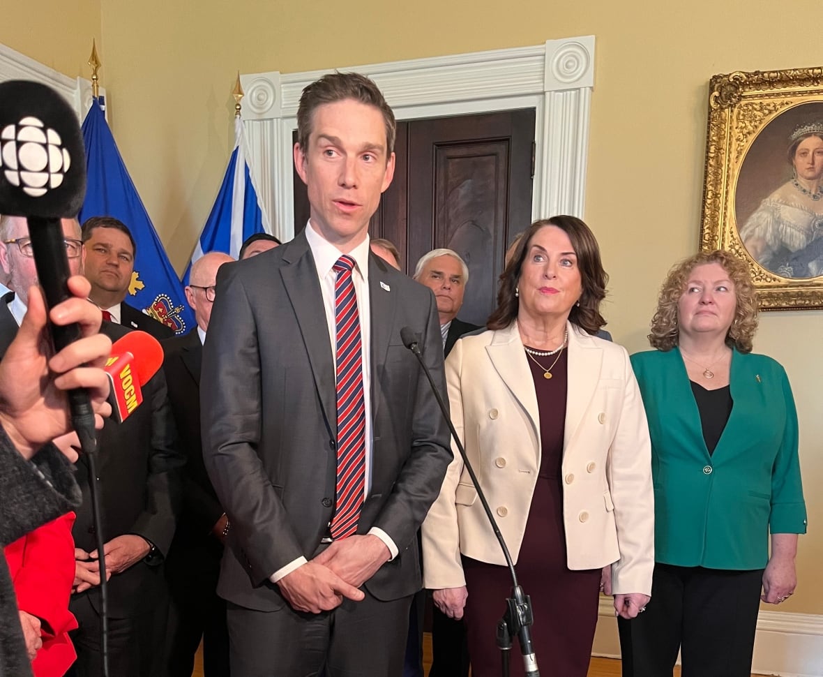 Man in grey suit and stripped tie standing in front of microphone, with group of people in suits behind him.