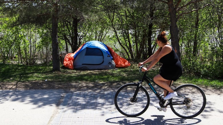 A cyclist riding on a sidewalk looks to their right and sees a blue tent pitched along a riverbank.