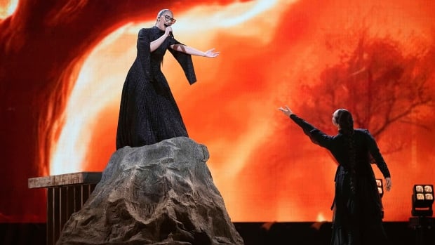 Female singer standing on a fake rock holding out her hand to another former on the stage below her.