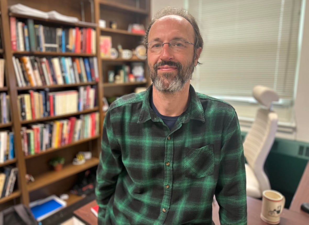 A man with brownish hair and a beard and a green plaid shirt leans on his desk with a book case to his right and a window behind him.