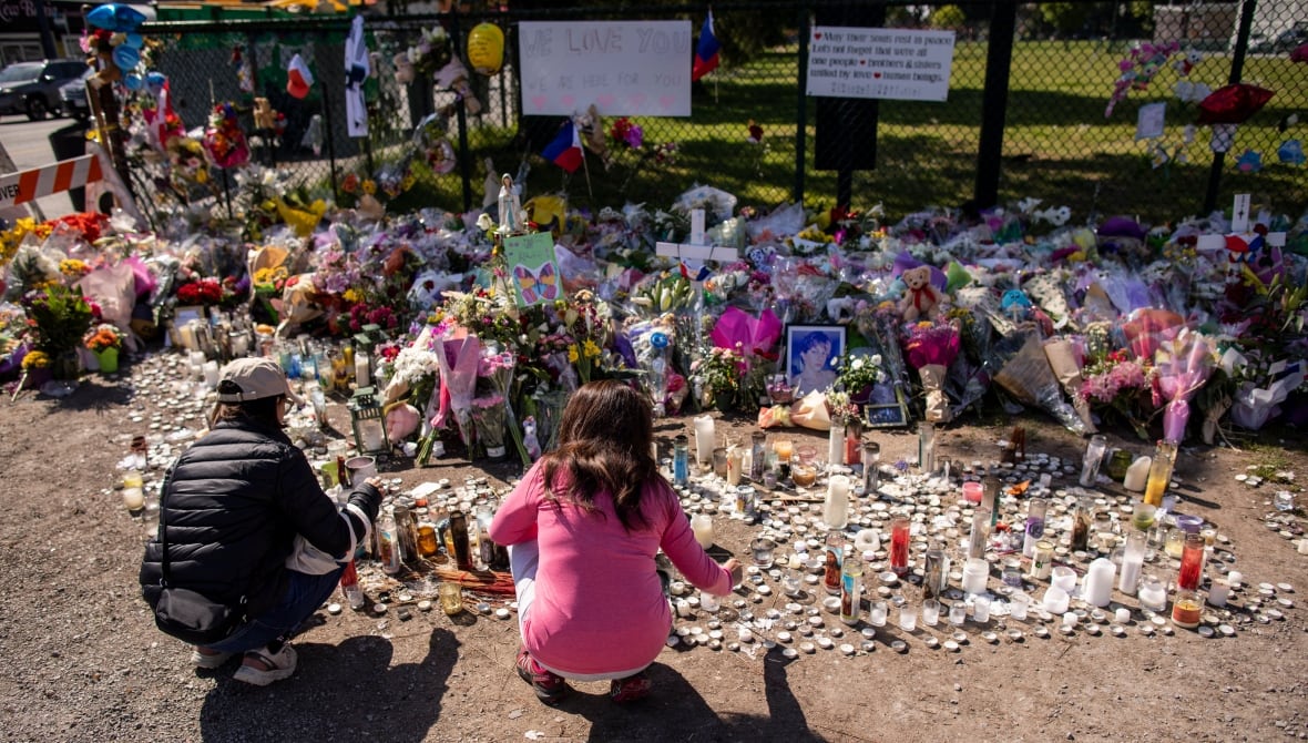 Two people kneel in front of candles, flowers and messages of sympathy.
