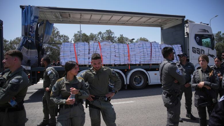 People successful  military-type uniform, men and women, basal   connected  pavement immoderate   metres distant  from a motortruck  that is loaded with bundles of material.