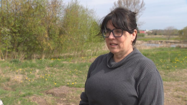 A woman with black hair, black glasses and a black shirt stands in front of bushes and there is some water in the background.
