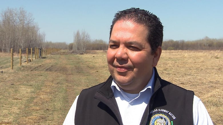 the chief (a man) stands on the grounds of the farm. fence posts meant to keep the animals in can be seen behind him