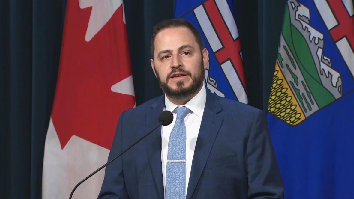 a man in a blue suit and blue tie stands at a microphone with alberta and canada flags behind