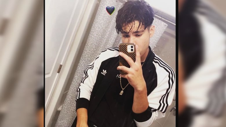 A man with short dark hair holds a cell phone in front of his face. He's wearing a black jacket with white sleeves and stands in front of a shower.