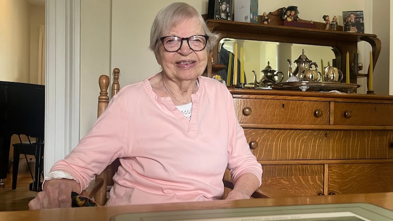 An elderly woman sits in a chair in her home