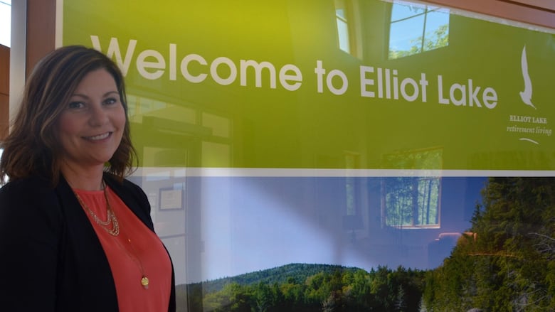 A woman stands in front of a sign that reads 'Welcome to Elliot Lake' 