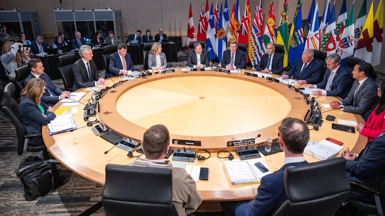 Politicians sit around a circular table with a row of flags in the background.