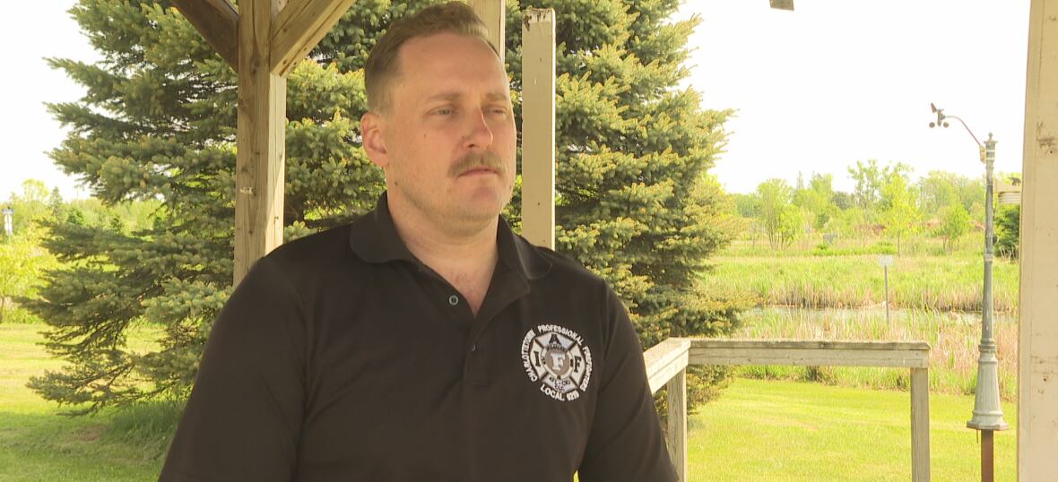 A man in a black shirt with a firefighter emblem stands underneath wooden structure with trees in the background. 