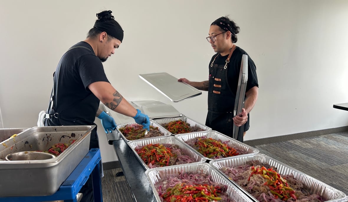 Two chefs are filling silver takeout containers with sautéed onions and peppers. 