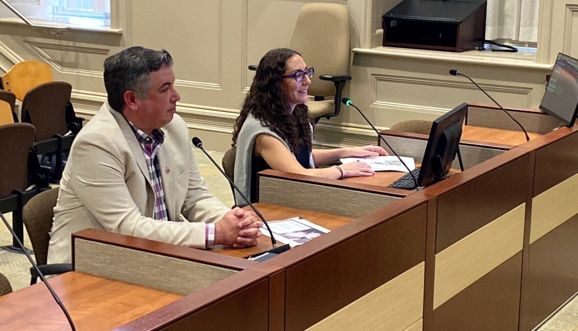 A white man with grey hair and a cream blazer sits at a desk with a microphone. Beside him is a white woman with long brown hair and glasses with a grey sweater.