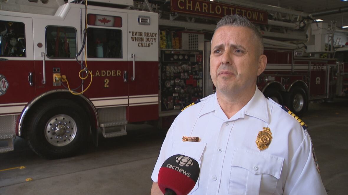 A man in a white shirt with a gold badge stands in front of a fire engine in a fire hall