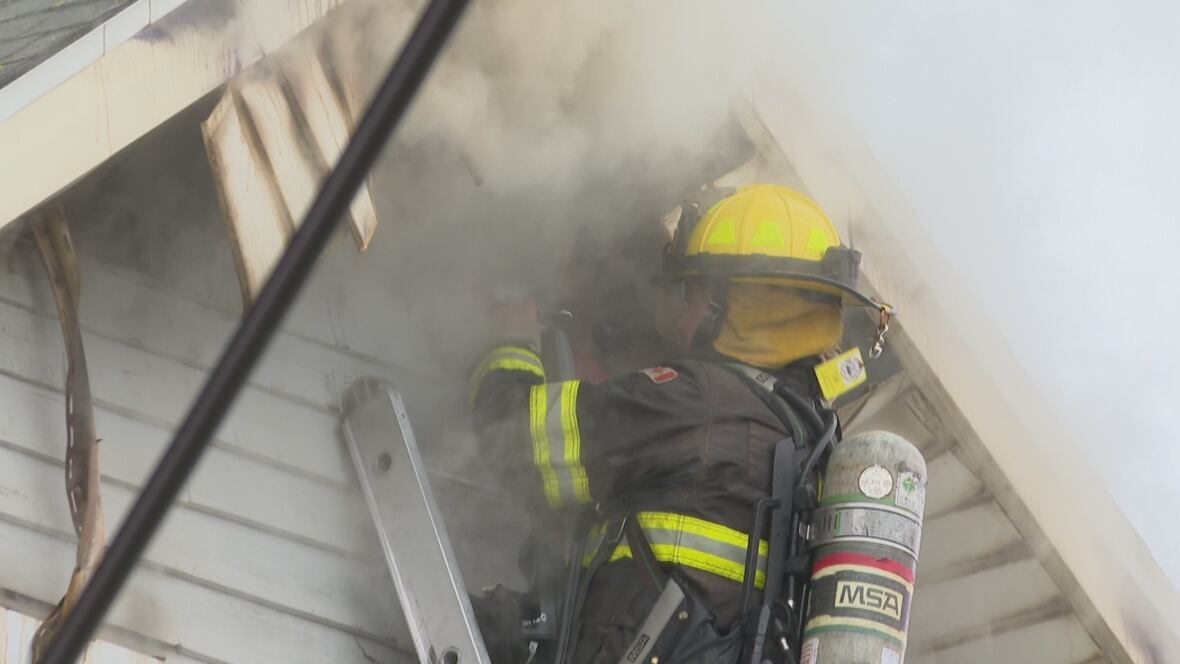 A firefighter is on a ladder and is underneath a overhanging rooftop, there is smoke all around them.