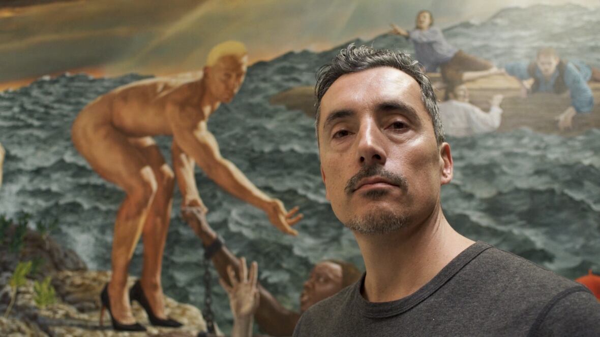 A man looks at the camera as he stands in front of a large painting featuring a naked man on a rocky short helping people from the water.