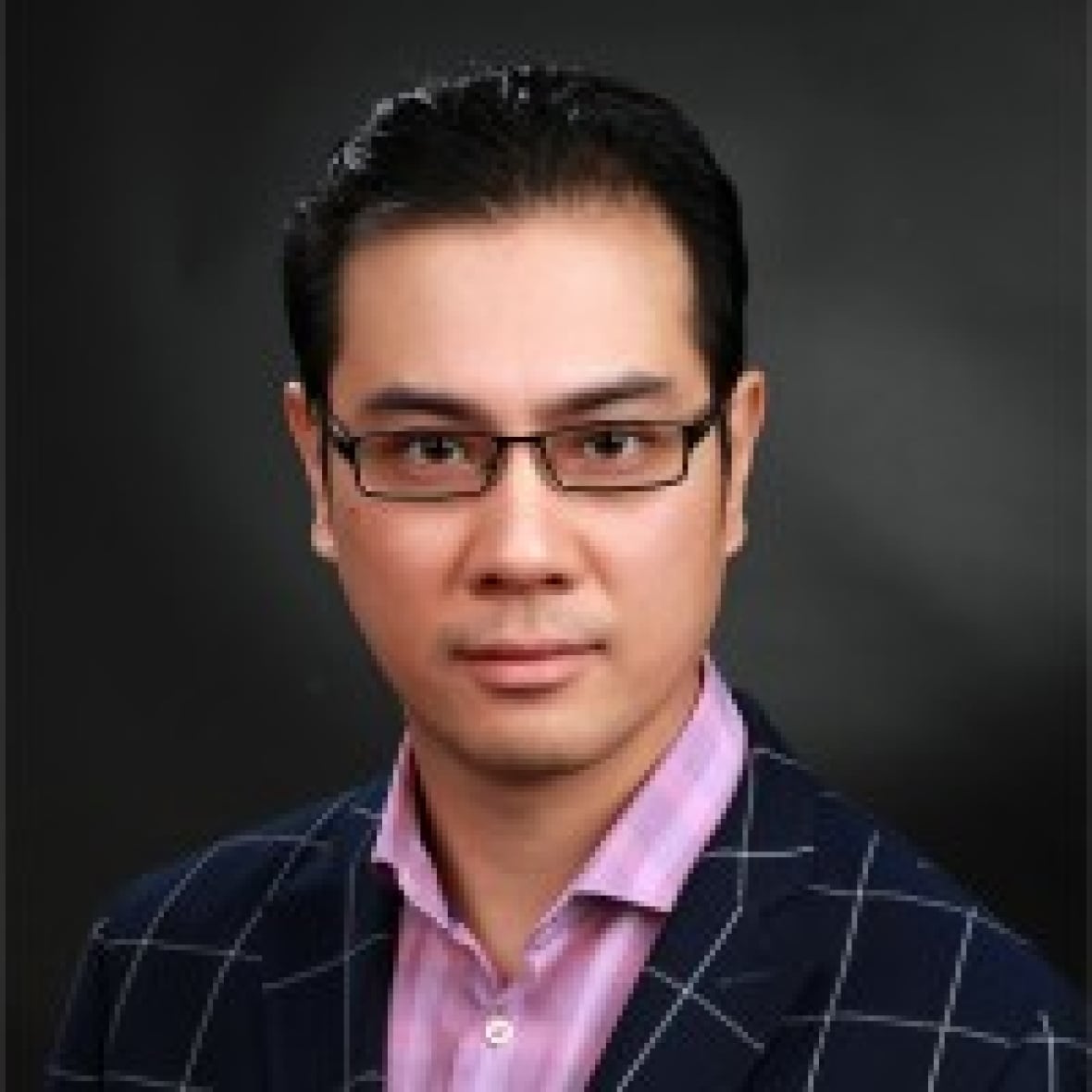 A man with dark slicked back hair and black square framed glasses in a professional head and shoulders portrait, wearing a dark blue jacket with a pattern of large squares bordered by thin white lines and a pink shirt.