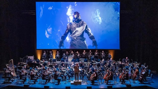 An orchestra preforms a piece with a photo of a robed figure visible.