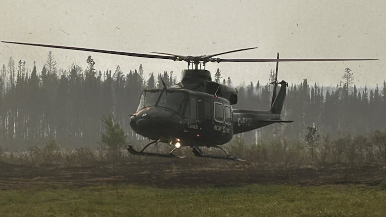 A military helicopter lifts off from the ground in a smoky area surrounded by trees.