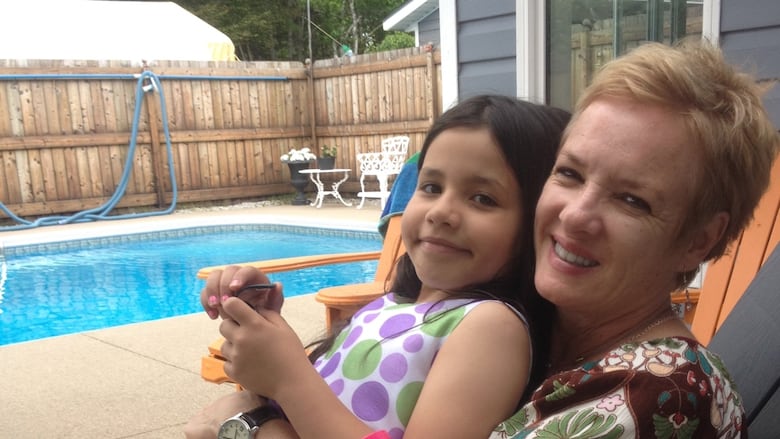 A woman with short hair sits on a lounge chair by a pool, with a black-haired girl seated on her lap and leaning back on her.
