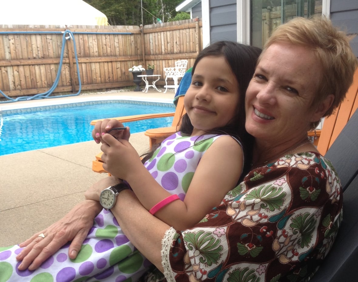 A woman with short hair sits on a lounge chair by a pool, with a black-haired girl seated on her lap and leaning back on her.