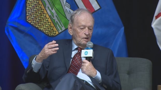 A man wearing a suit is pictured sitting in a chair and talking into a microphone in front of an Alberta flag.