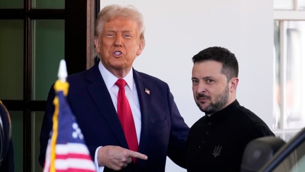 A man in a suit points to another man. They are side by side and near an American flag.