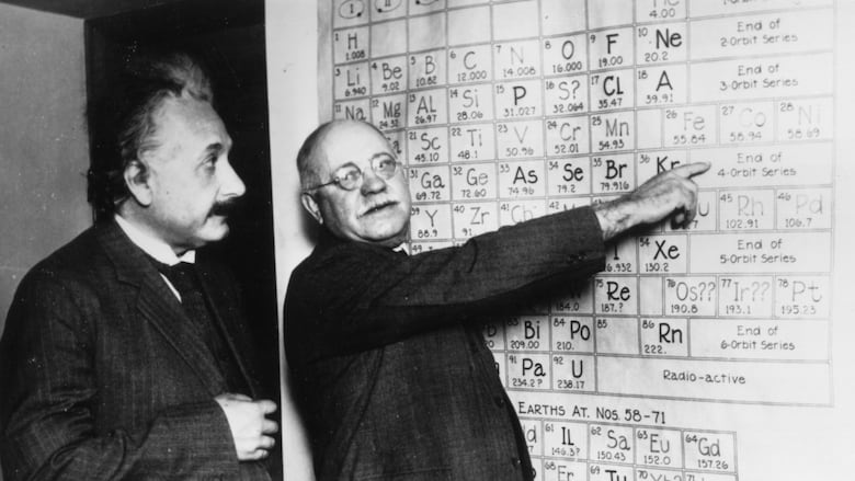 Two men stand at a whiteboard, one points at a periodic table of elements.