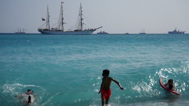 Children play in waves on beach as boats sail in the background