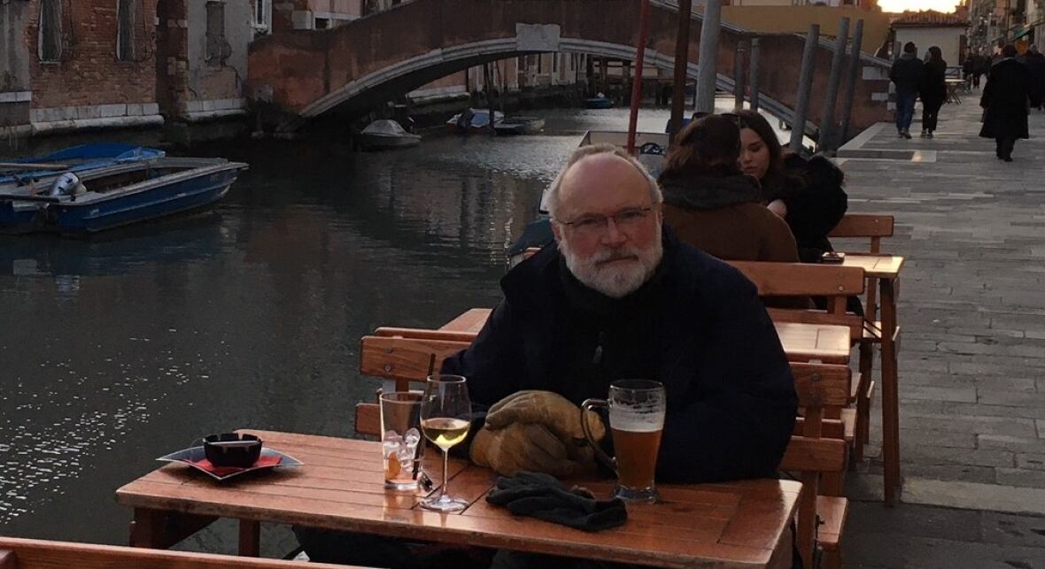 A man sitting at a table alongside a canal with a row of tall brick buildings in the background and a small boat in the water. 