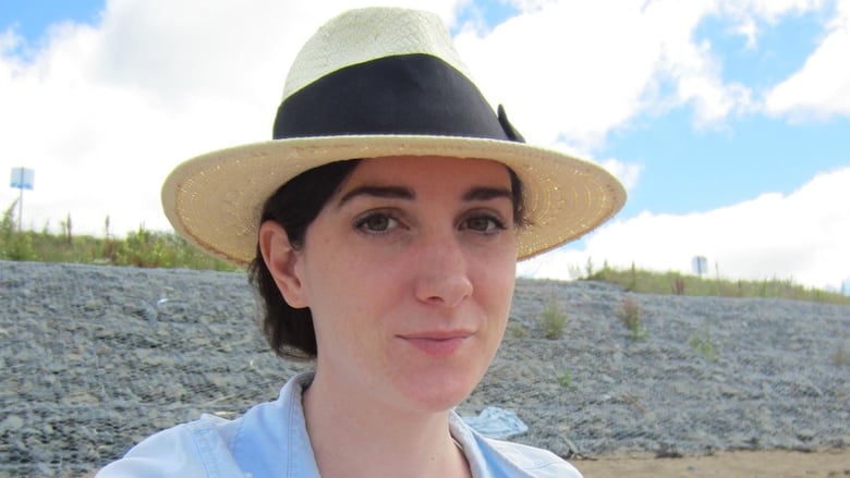 A woman sitting on a beach with a straw hat on and a light blue dress shirt with rolled up sleeves.
