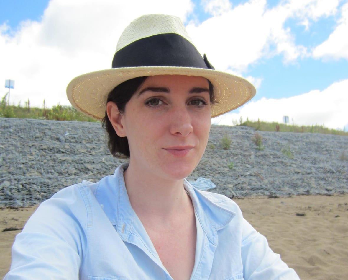 A woman sitting on a beach with a straw hat on and a light blue dress shirt with rolled up sleeves.