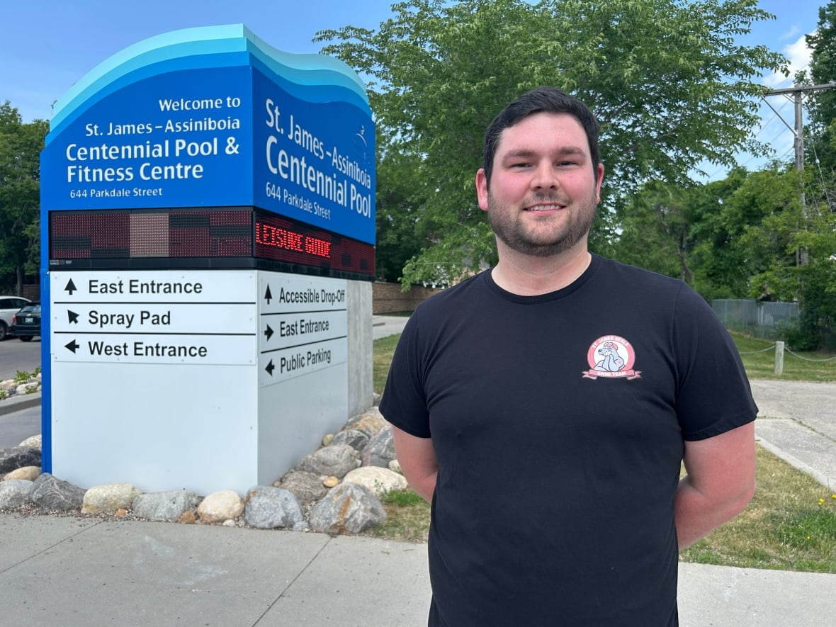 Man with dark hair and dark eyes wears a navy blue t-shirt with a red and white chest emblem. He stands next to a sign for a City of Winnipeg pool. 