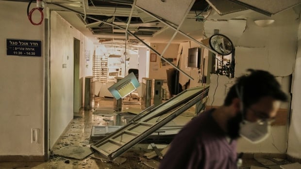A person wearing a mask over their mouth is shown in the foreground inside a building with damage and doors and debris on the ground.