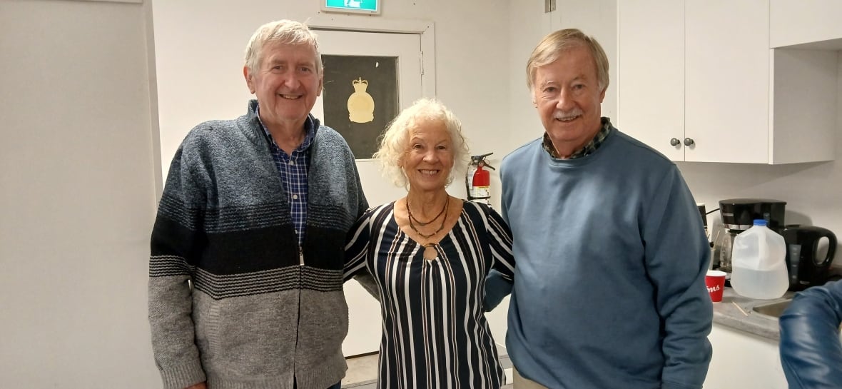 Two older men and an older woman smile at the camera with their arms behind each other.