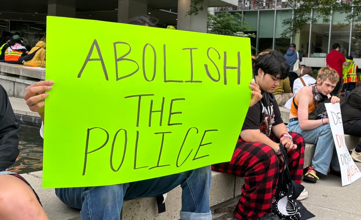 People sitting on a ledge, someone holds a sign that reads "abolish the police."