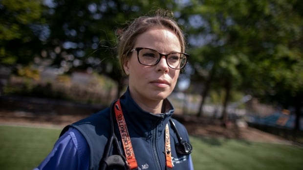 A woman with short brown hair wearing glasses, a blue shirt and vest, with an identification lanyard around her neck, stands outside in a green, treed area.