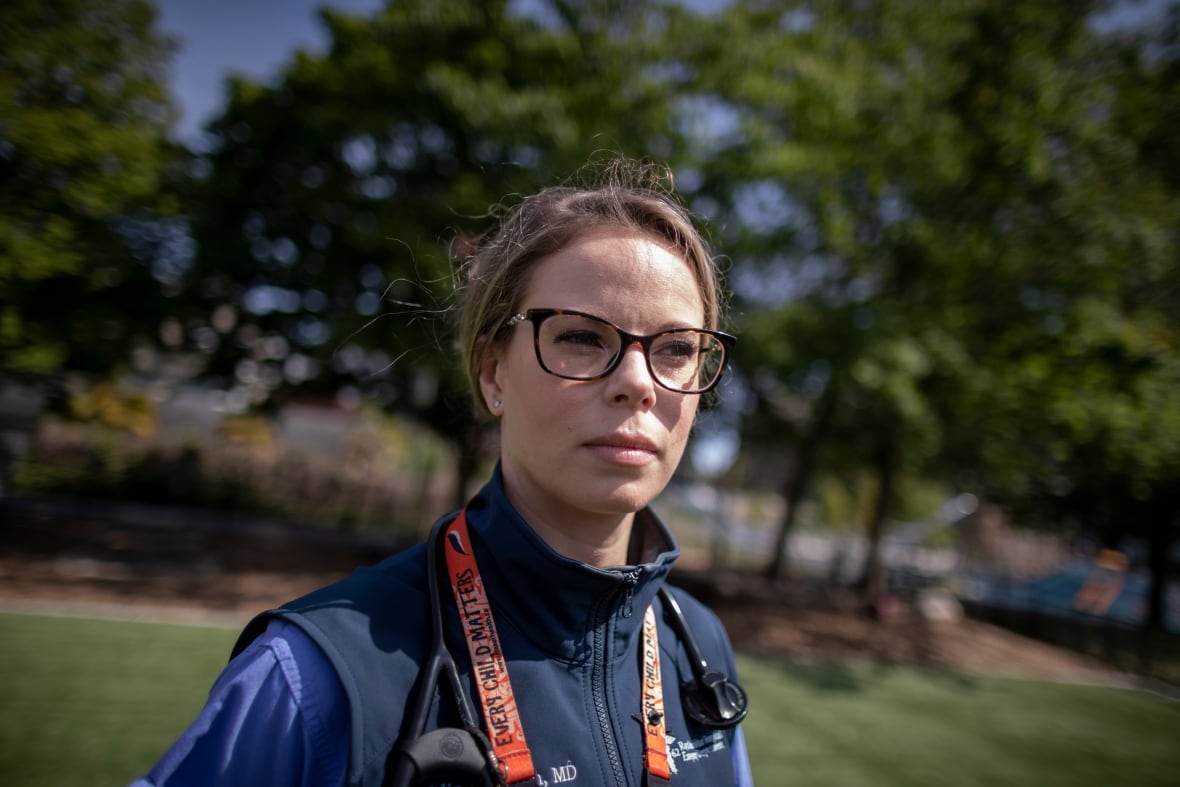 A woman with short brown hair wearing glasses, a blue shirt and vest, with an identification lanyard around her neck, stands outside in a green, treed area.