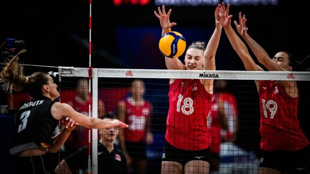 Two female volleyball players representing Canada jump up to block the ball at the net.