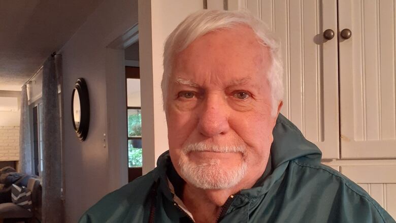 A smiling man with white hair and a goatee in a gray rain jacket sitting in a living room. 