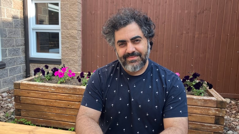 A man sitting at a table in a blue t-shirt. He has a greying beard and black hair and sits in front of a flower bed. 