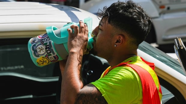 Wearing a yellow T-shirt and safety vest, a worker drinks from a giant canister near a vehicle, amid a heat wave.