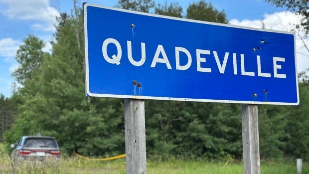 Road sign that says "Quadeville" with an SUV in the background.