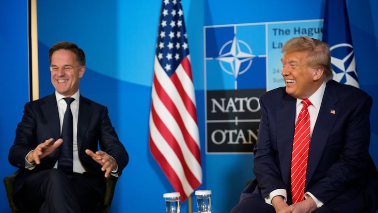 Two cleanshaven men in suit and tie sit, separated by just a small table.