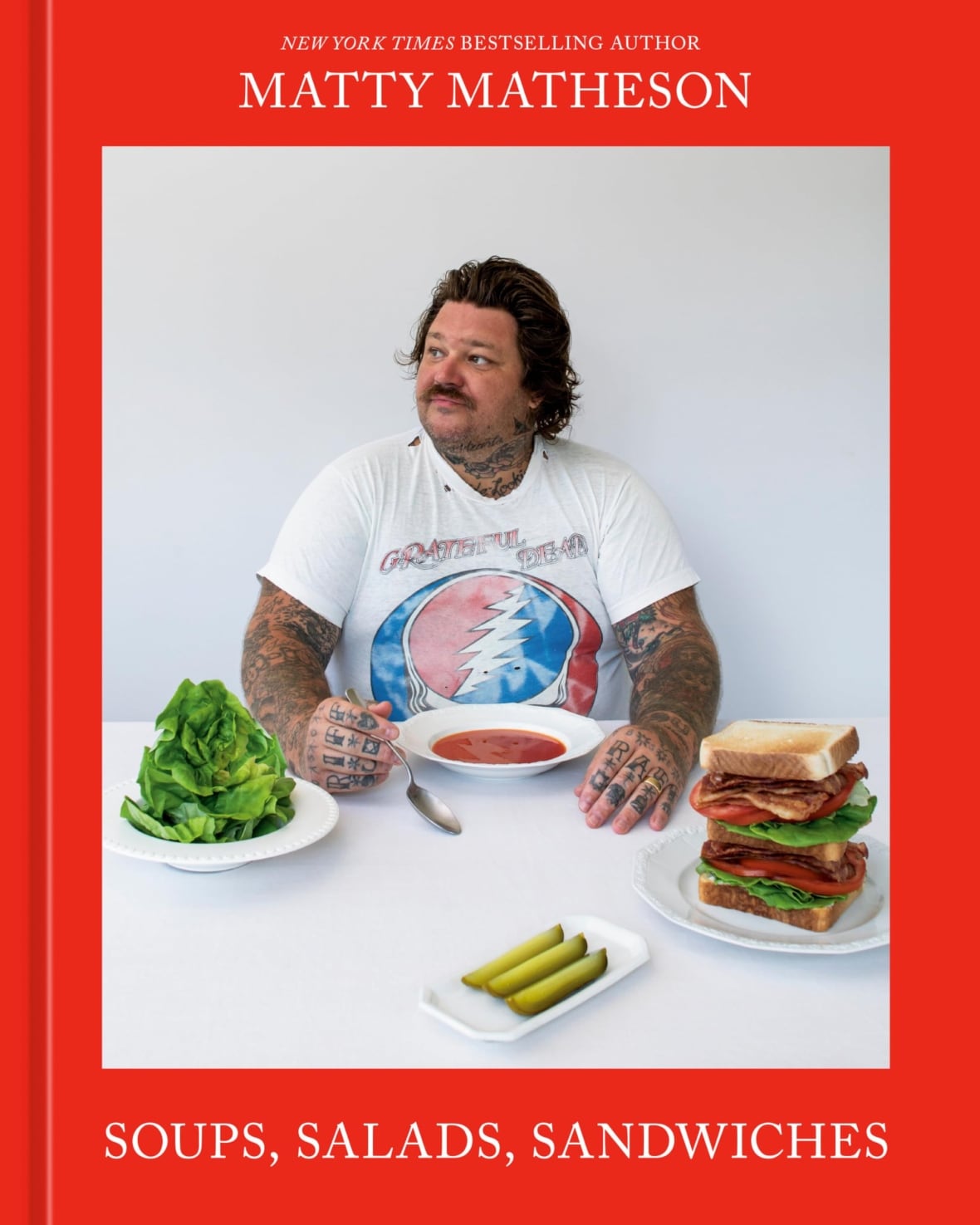 A book cover of a man with long hair and tattoos sitting at a table with a salad, sandwiches and a bowl of soup.